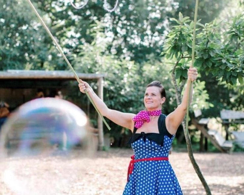 A smiling woman holds two sticks aloft as bubbles form between them, hanging from strings